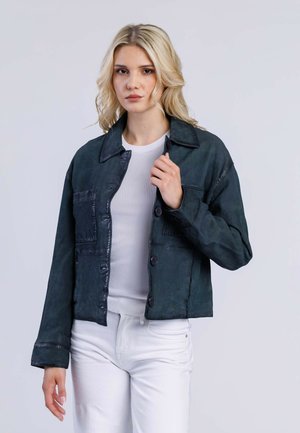 Veste en denim vert foncé avec col, poches avant et fermeture à boutons. Présente une finition texturée et une coupe décontractée. Top blanc à côtes en dessous.