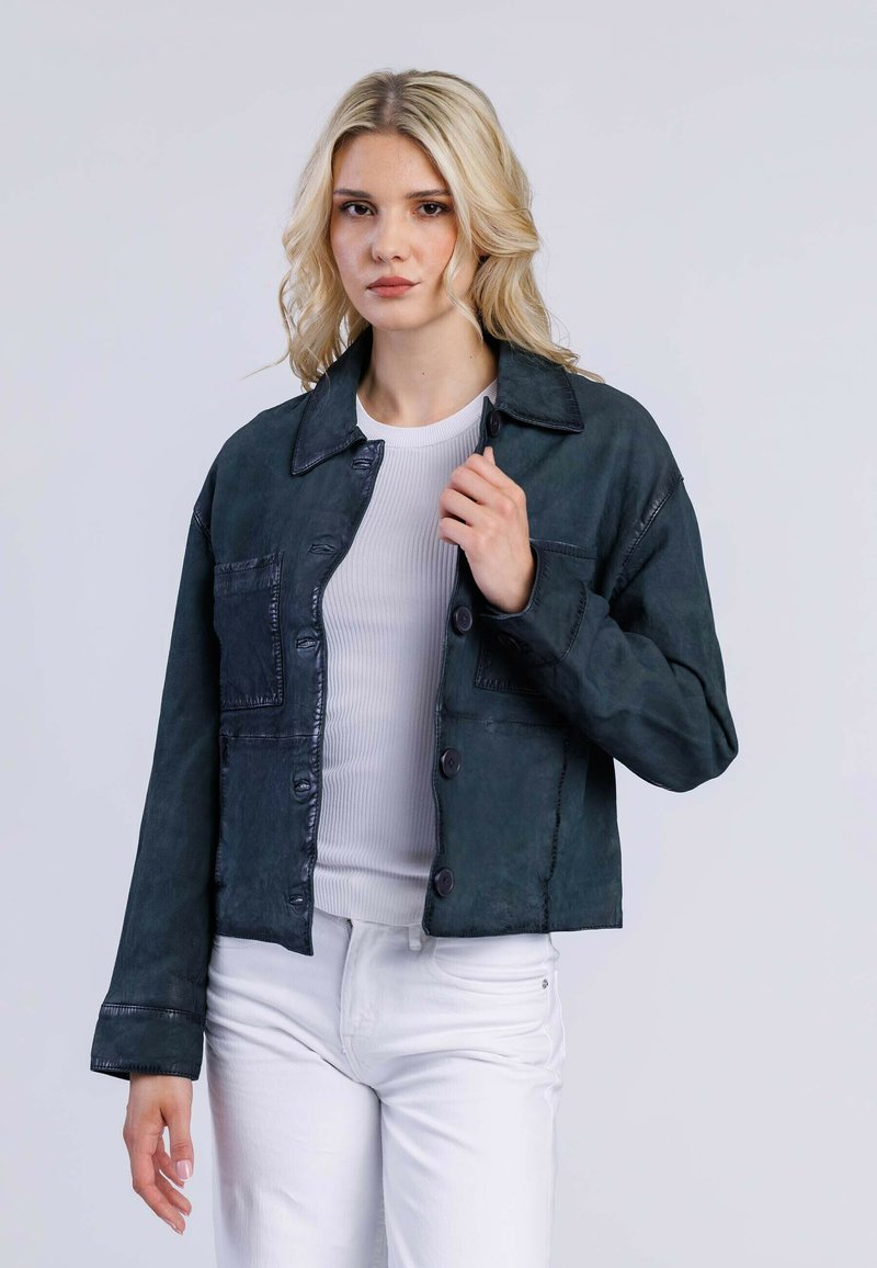 Donkergroene denimjack met een kraag, voorzakken en knoopsluiting. Heeft een gestructureerde afwerking en een relaxte pasvorm. Witte geribbelde top eronder.