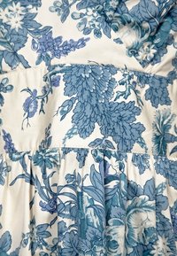 Tissu fleuri bleu et blanc avec des motifs de feuilles et de fleurs, montrant des coutures cousues et des plis froncés.