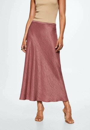 Vrouw die een glanzende mauve enkellange rok draagt, beige mouwloze top en beige sandalen met hoge hak en bandjes, staande tegen een witte achtergrond.