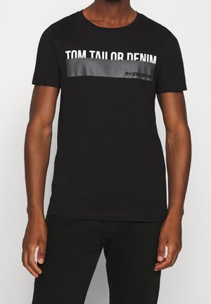 Mężczyzna ubrany w czarną koszulkę z krótkim rękawem z napisem „TOM TAILOR DENIM” i szarym poziomym paskiem na wysokości klatki piersiowej.
