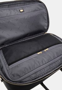 Intérieur d'un sac en tissu noir avec une doublure en satin gris présentant un motif répétitif "GUESS", une poche en maille et des accents en métal doré.