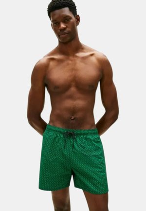 Hombre musculoso con piel oscura y cabello corto y rizado, vestido con pantalones cortos de baño verdes con estampado, de pie con las manos detrás de la espalda sobre fondo blanco.