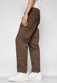 Brown cargo pants with multiple side pockets, straight cut, made from sturdy fabric, paired with white sneakers in a side profile view.