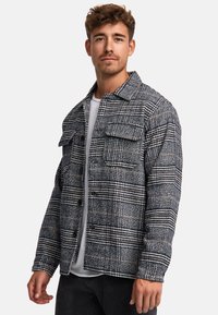 Plaid shirt jacket in black, white, and brown hues; features button-down front, two chest pockets, and textured fabric.