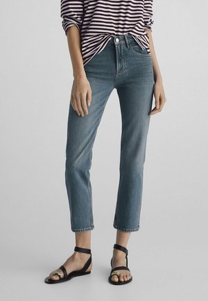 Person trägt blaue, gekürzte Jeans, schwarze Sandalen mit Knöchelriemen und ein lila-weiß gestreiftes Langarmshirt, steht vor einem einfarbigen Hintergrund.