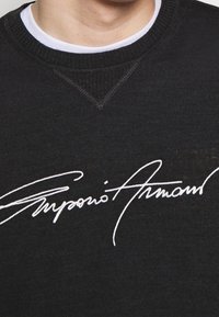 Black sweatshirt with a textured knit, featuring a white embroidered signature design across the chest. Round neckline and cuffed sleeves.