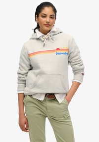 Felpa grigia chiara con cappuccio e stringhe, tasca a marsupio e logo grafico colorato con strisce orizzontali. Indossata sopra una maglietta bianca e pantaloni verdi.