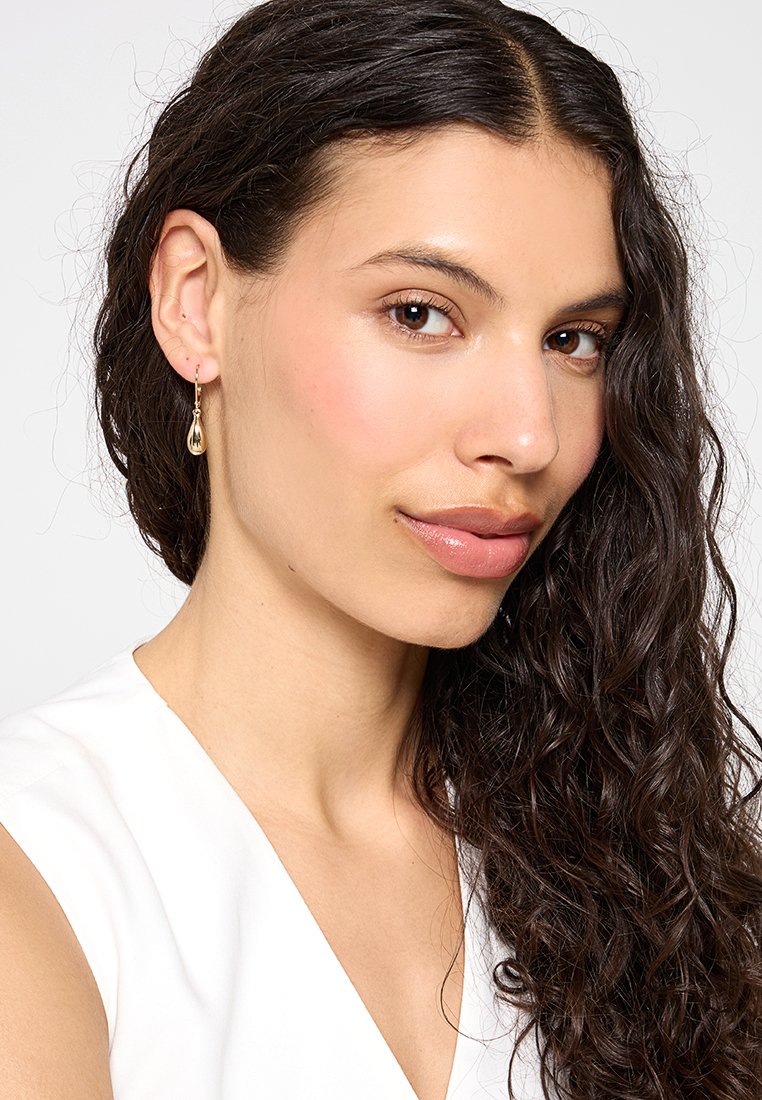 Mujer con cabello largo y rizado de color oscuro, vistiendo una blusa sin mangas blanca y aretes en forma de lágrima dorados, mirando ligeramente de lado.
