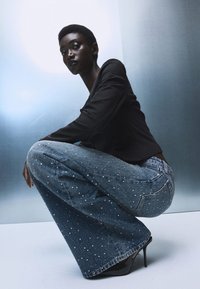 Jeans a zampa d'elefante in denim azzurro chiaro con macchie bianche, abbinati a un top aderente nero. La modella è accovacciata, mettendo in evidenza la gamba larga e la texture dei jeans.