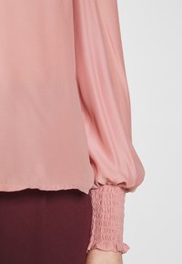 Blouse rose à manches longues avec poignet élastique froncé et léger détail de volant, portée avec un pantalon bordeaux foncé sur un fond clair.