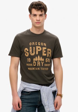 Camiseta de color verde oliva oscuro con un gráfico impreso en beige, que presenta "OREGON SUPER DRY SHOP" y diseños de árboles, usada con una camisa a rayas azul anudada en la cintura.