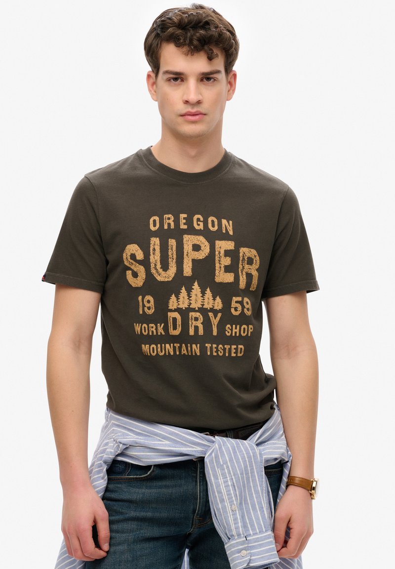 Camiseta de color verde oliva oscuro con un gráfico impreso en beige, que presenta "OREGON SUPER DRY SHOP" y diseños de árboles, usada con una camisa a rayas azul anudada en la cintura.