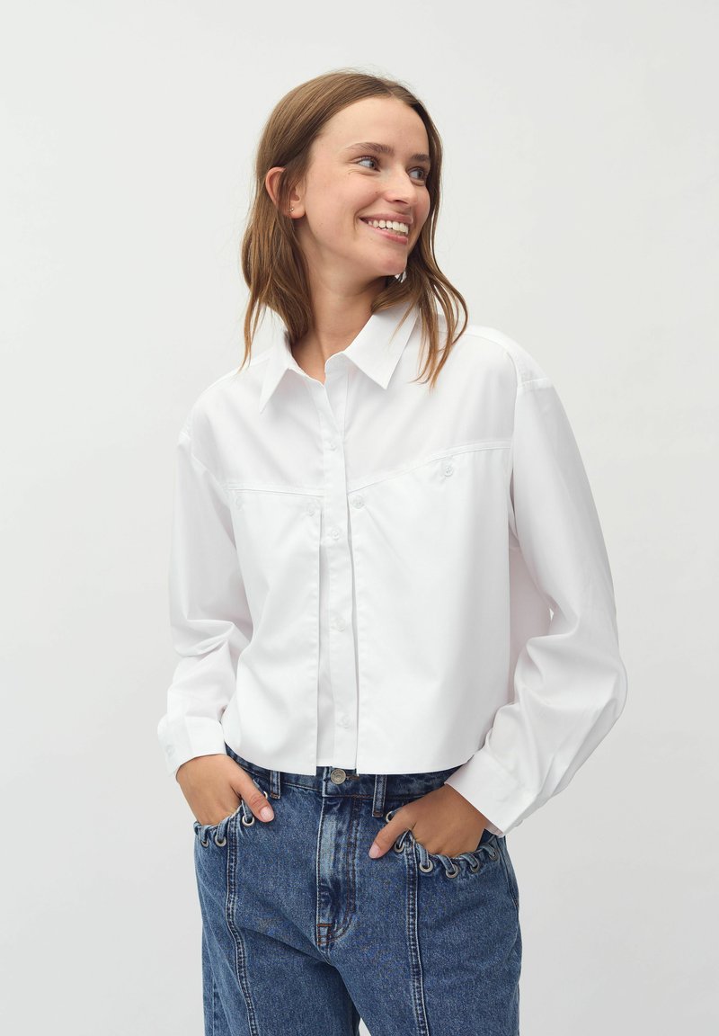 White button-up shirt with a collar and long sleeves. The fabric is smooth, paired with high-waisted blue jeans featuring unique stitching details.