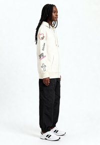 Kremowa bluza z kapturem z nadrukami graficznymi, w tym sercem i tekstem. Połączona z czarnymi spodniami cargo i białymi trampkami z czarnymi akcentami.