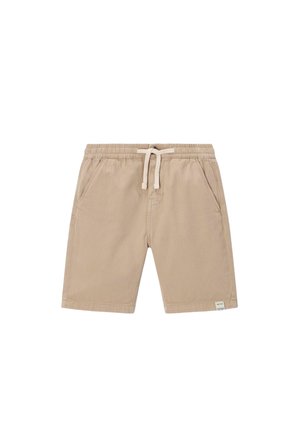 Kind met krullend haar dat een lichtblauwe pet, crèmekleurige trui, beige korte broek, gebreide sokken met patroon en beige sneakers draagt, staat met handen in de zakken.
