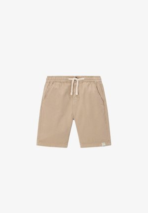 Kind met krullend haar dat een lichtblauwe pet, crèmekleurige trui, beige korte broek, gebreide sokken met patroon en beige sneakers draagt, staat met handen in de zakken.