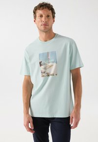 Camiseta de algodón de color azul claro con un gráfico rectangular que presenta el texto "SALSA JEANS", mostrando imágenes de playa y colores suaves.