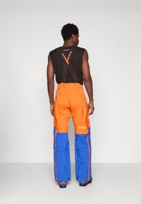 Άνδρας που φοράει πορτοκαλί και μπλε παντελόνια εξωτερικού χώρου με τιράντες και μαύρη αμάνικη μπλούζα, στέκεται μπροστά σε απλή ανοιχτόχρωμη φόντο.