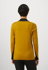 Long-sleeved top in mustard-yellow fabric with black collar and side accents, featuring a smooth texture and form-fitting design.