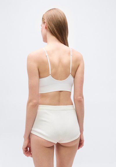 Γυναίκα με μακριά μαλλιά στέκεται γυρισμένη πλάτη, φοράει λευκά ριμπ ψηλόμεσα σορτς και λευκό σουτιέν με λεπτές ρυθμιζόμενες τιράντες.