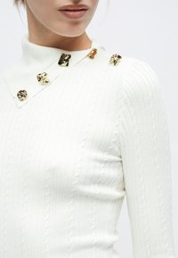 Λευκό ριγωτό πουλόβερ με ψηλό γιακά και χρυσές διακοσμητικές λεπτομέρειες στον ώμο. Υφή υφάσματος με πλεκτό μοτίβο καλωδίων.