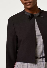 Blazer noir court avec un col en satin, orné d'un détail en forme de nœud. Porté sur une robe à carreaux gris avec des lignes discrètes et une taille cintrée.