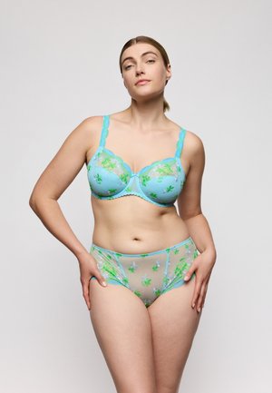 Vrouw staat met handen in de zij, draagt een lichtblauw doorschijnend lingeriesetje met groene en witte bloemrijke borduursels, tegen een effen lichte achtergrond.