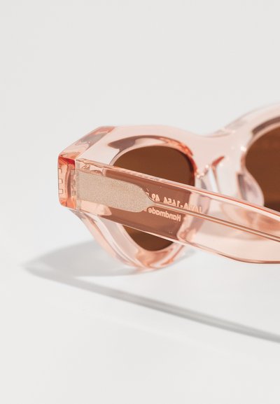Ochelari de soare de culoare piersici, cu lentile rotunde, rame transparente groase și o textură netedă. Branding notabil pe braț.