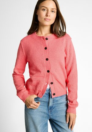 Woman wearing a coral pink buttoned cardigan over a white top, paired with light blue denim jeans, posing with one hand in pocket.