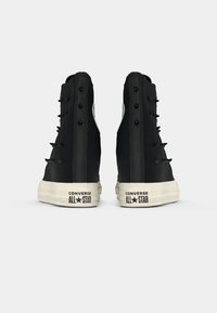 Čierne vysoké tenisky Converse All Star s bielymi podrážkami, pohľad zozadu na jednoduchom svetlom pozadí.