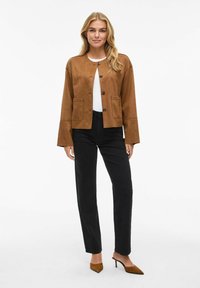 Veste cropped en suede marron avec deux poches avant, fermeture à boutons et manches longues, associée à un pantalon noir et des chaussures à bout pointu.