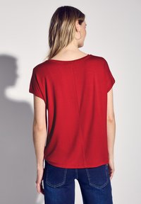 Camiseta corta de manga corta roja con un ajuste relajado, textura suave y cuello redondeado, que presenta una costura central en la parte trasera. Combinada con jeans azules.