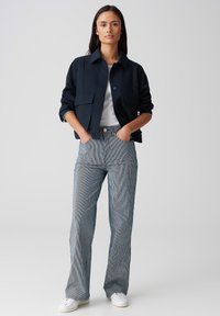 Veste cropped marine avec deux poches avant, portée sur une chemise blanche. Pantalon taille haute à rayures bleues et blanches, associé à des baskets blanches.