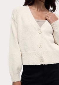 Cardigan en tricot de couleur crème avec un col en V, trois boutons et un tissu texturé. Il présente des manches longues légèrement bouffantes et un design cropped.