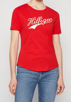 Osoba ubrana w czerwoną koszulkę z krótkim rękawem z białym tekstem "Hilfiger" i niebieskie dżinsy, przedstawiona od szyi do bioder na jednolitym tle.