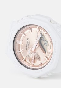 Montre G-Shock blanche avec un cadran métallisé doré rose, affichage numérique, lunette texturée et repères horaires proéminents. Étanche jusqu'à 20BAR.