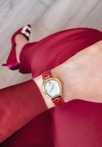 Złoto-tonowy zegarek na nadgarstek z białą tarczą i czerwonym paskiem z krokodylim wzorem, noszony na nadgarstku, obok bordowej sukienki i pasujących szpilek.