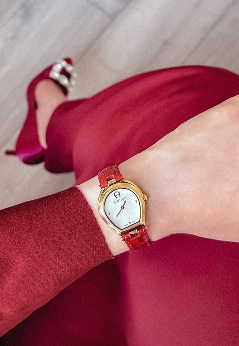 Złoto-tonowy zegarek na nadgarstek z białą tarczą i czerwonym paskiem z krokodylim wzorem, noszony na nadgarstku, obok bordowej sukienki i pasujących szpilek.