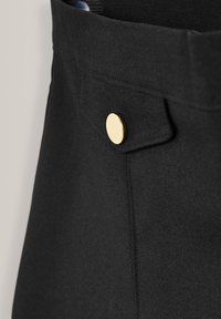 Vêtement en tissu noir avec un gros plan sur une poche à rabat fermée par un bouton rond en or.