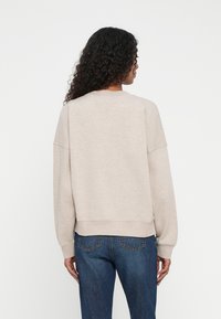 Mujer con cabello rizado, vistiendo un sudadera de manga larga beige y jeans azules, de pie de espaldas contra un fondo blanco liso.