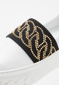 Biały but wsuwany z czarną elastyczną taśmą ozdobioną wzorem z łańcuszka z złotych kryształków, biała teksturowana podeszwa zapewniająca przyczepność i stabilność.