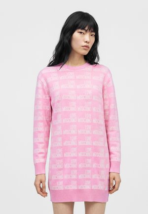 Femme aux longs cheveux noirs portant une robe rose à manches longues ornée du texte "LOVE MOSCHINO" sur un fond uni.