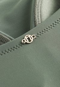 Zbliżenie zielonej tkaniny z przeszyciem w zygzak i małym okrągłym metalowym logo przymocowanym blisko szwu.