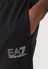 Pantaloni da jogging neri realizzati in tessuto morbido con vestibilità slim. Presentano una tasca laterale e un logo bianco "EA7 Emporio Armani" sulla coscia sinistra.