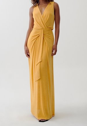 Mujer con un vestido maxi amarillo mostaza sin mangas, con un diseño cruzado en la parte delantera y un detalle de lazo largo en la cintura, posando sobre un fondo claro.
