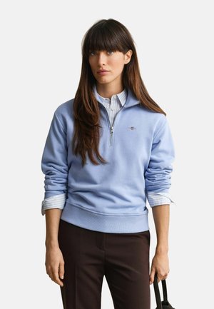 Frau mit langen braunen Haaren, die einen hellblauen Viertel-Zip-Sweatshirt über einem gestreiften Hemd und dunklen Hosen trägt, hält einen schwarzen Taschengriff.