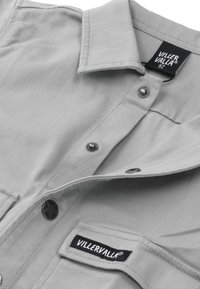 Jasnoszara koszula z zapięciem na guziki, z czarnymi metalowymi guzikami, kieszenią na piersi oraz metką marki wszytą w kieszeni.