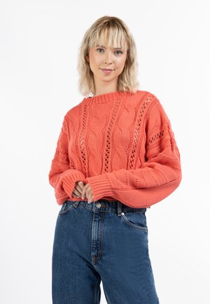 Pull en laine corail tricoté avec des motifs en câble et des détails en trame ouverte. Manches longues et poignets côtelés, associé à un jean en denim taille haute.