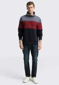 Grijze, rode en marineblauwe gestreepte pullovertrui met een hoge kraag en knoopdetails. Gecombineerd met donkere jeans en witte sneakers.